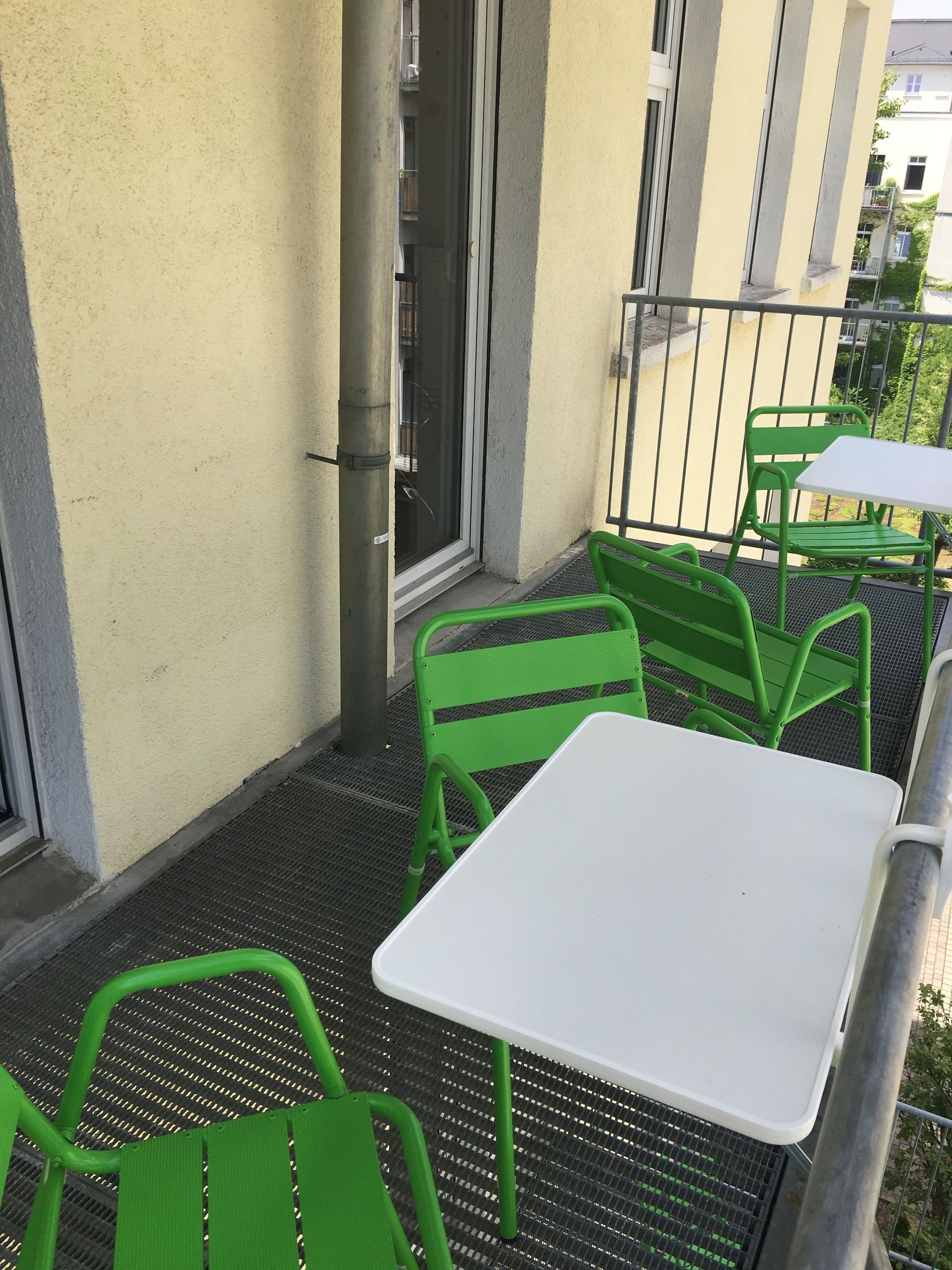 Un balcón con sillas de metal verde y una mesa blanca, baranda metálica y un edificio al fondo.