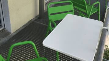 Un balcón con sillas de metal verde y una mesa blanca, baranda metálica y un edificio al fondo.