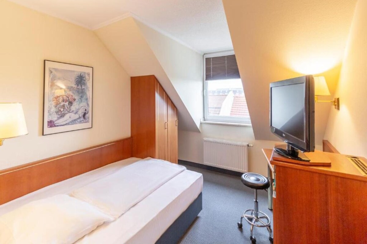 Habitación de hotel con cama, escritorio con computadora, televisor, ventana con persianas y radiador.