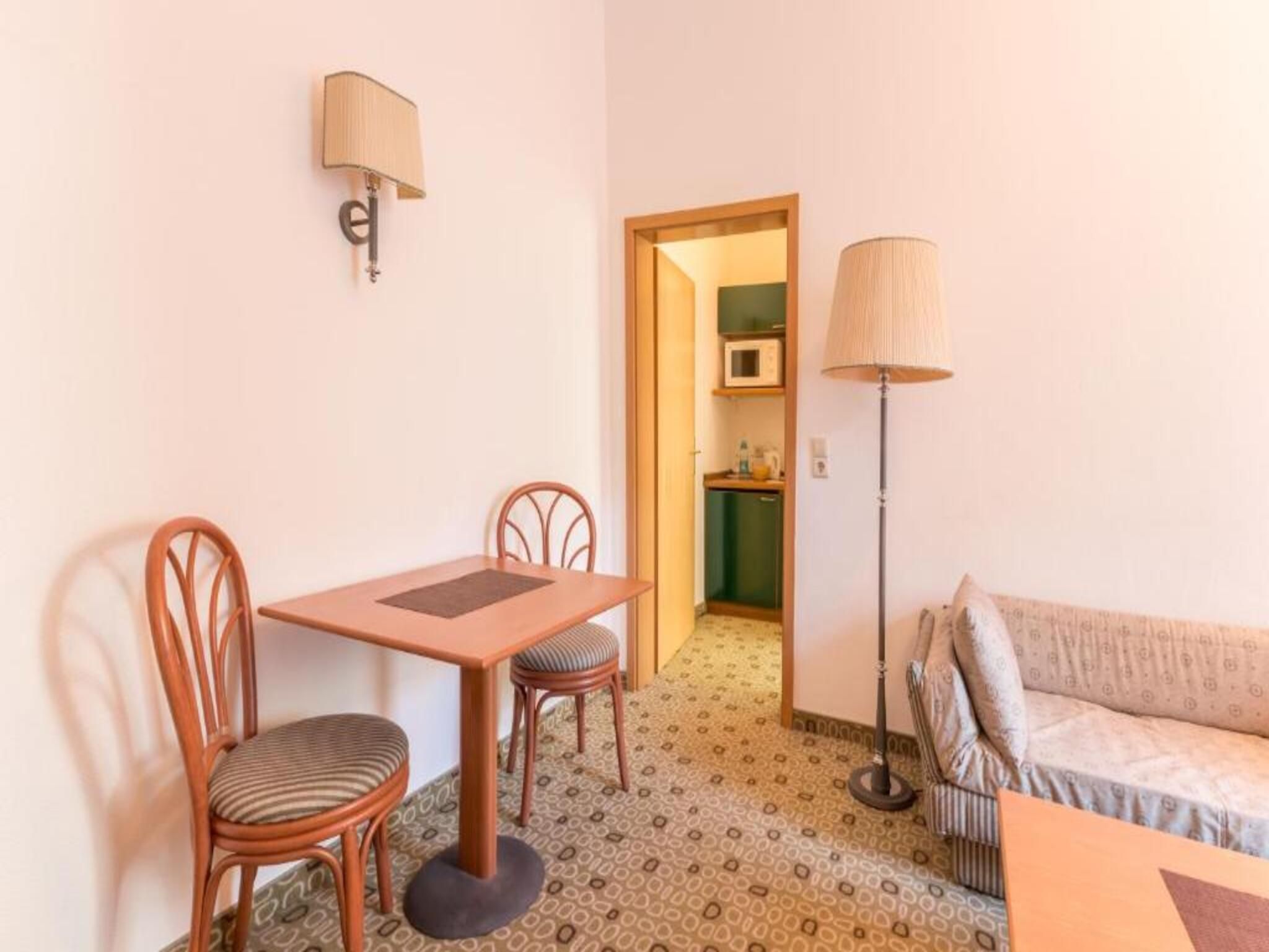 Una habitación pequeña con una mesa, dos sillas, un sofá, una lámpara y un microondas al fondo.