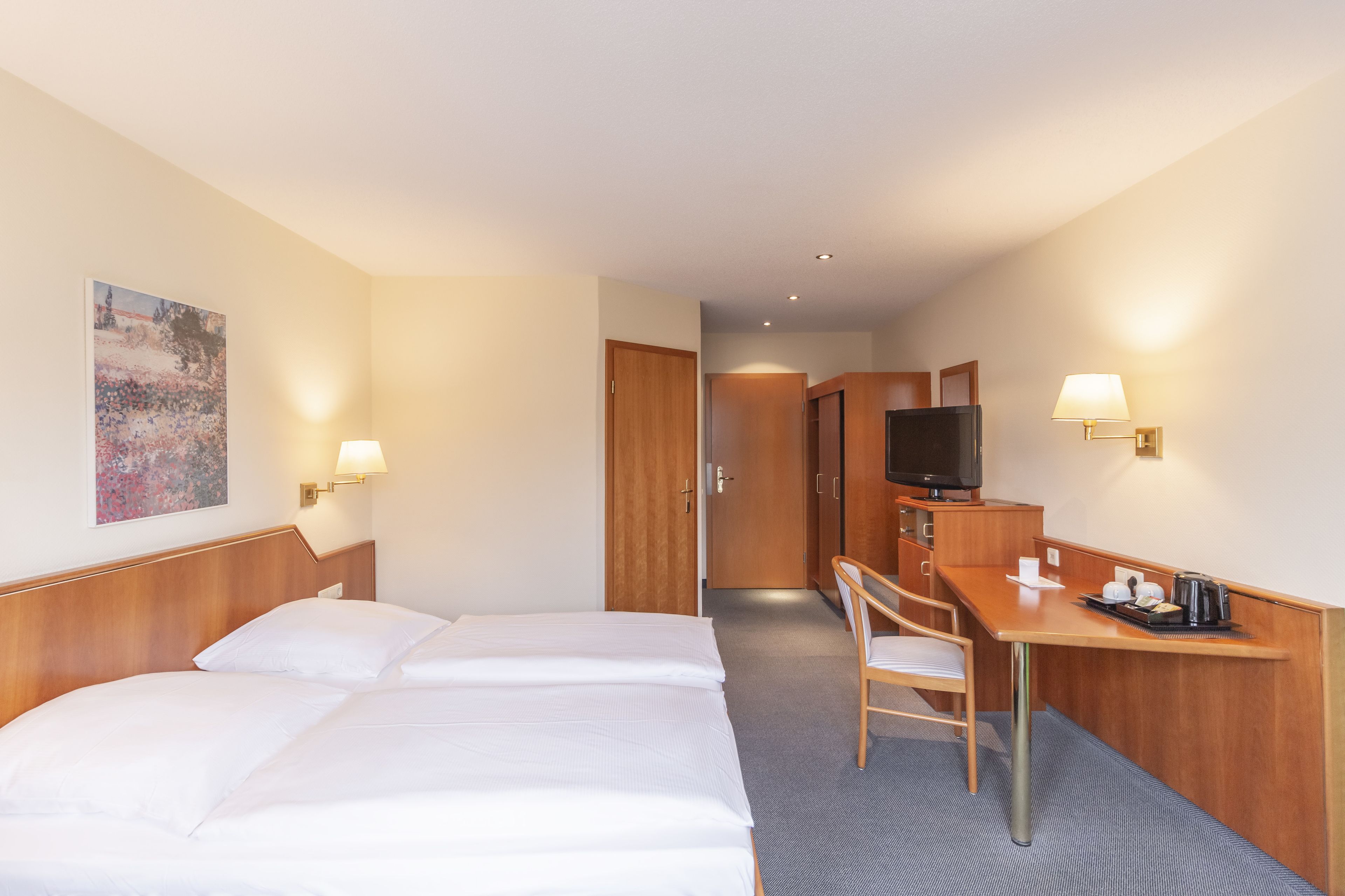 A hotel room with a bed, a desk, a chair, a television, and a painting on the wall.