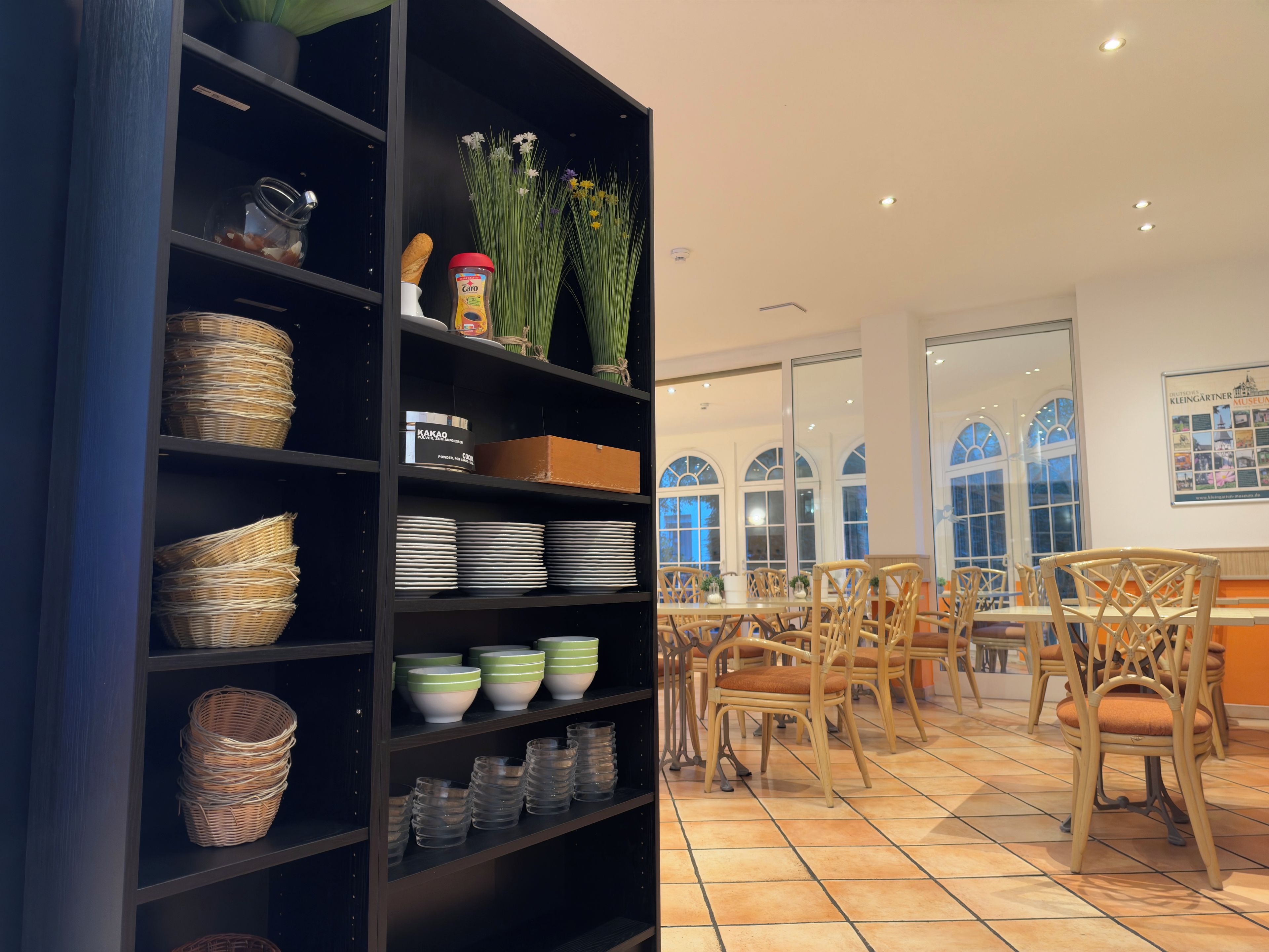 Ein Zimmer mit einer schwarzen Regaleinheit, die Körbe, Teller und Gläser enthält. Es gibt Korbstühle und einen Tisch mit einem Blumenstrauß.