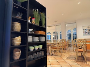 Una habitación con un mueble de estantería negro que contiene cestas, platos y frascos. Hay sillas de mimbre y una mesa con un jarrón de flores.