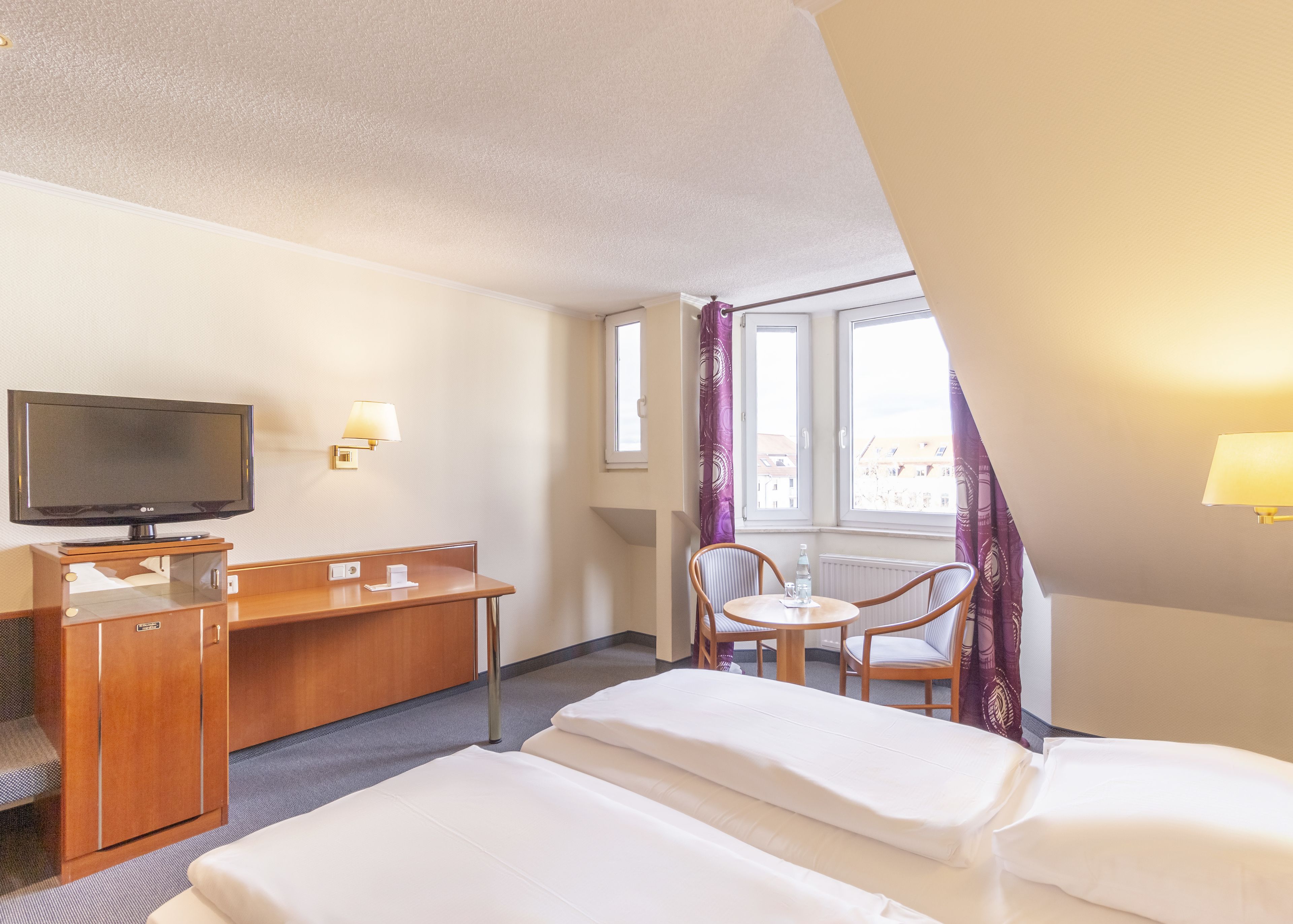 A hotel room with a bed, a desk with a TV, a chair, and a window with curtains.