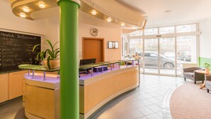 Un espace d’accueil doté d’un comptoir incurvé, d’un pilier vert, d’un tableau noir avec du texte, d’une horloge et d’une vue sur les voitures garées, visible à travers de grandes fenêtres.