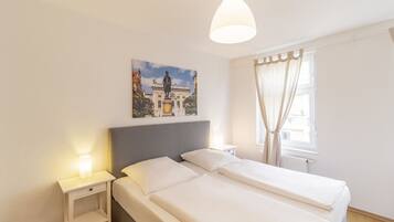 A hotel room with a bed, a nightstand, a wall-mounted painting, a window with curtains, and a hanging light fixture.