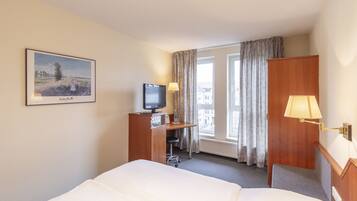 A hotel room with a bed, a desk with a television, a lamp, and a window with curtains.