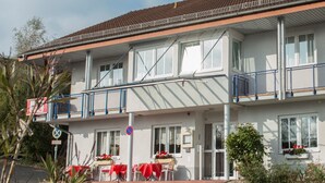 Un bâtiment à deux étages doté d’un balcon, d’un espace extérieur aménagé avec des tables et des chaises rouges, et de jardinières sur les rebords des fenêtres.