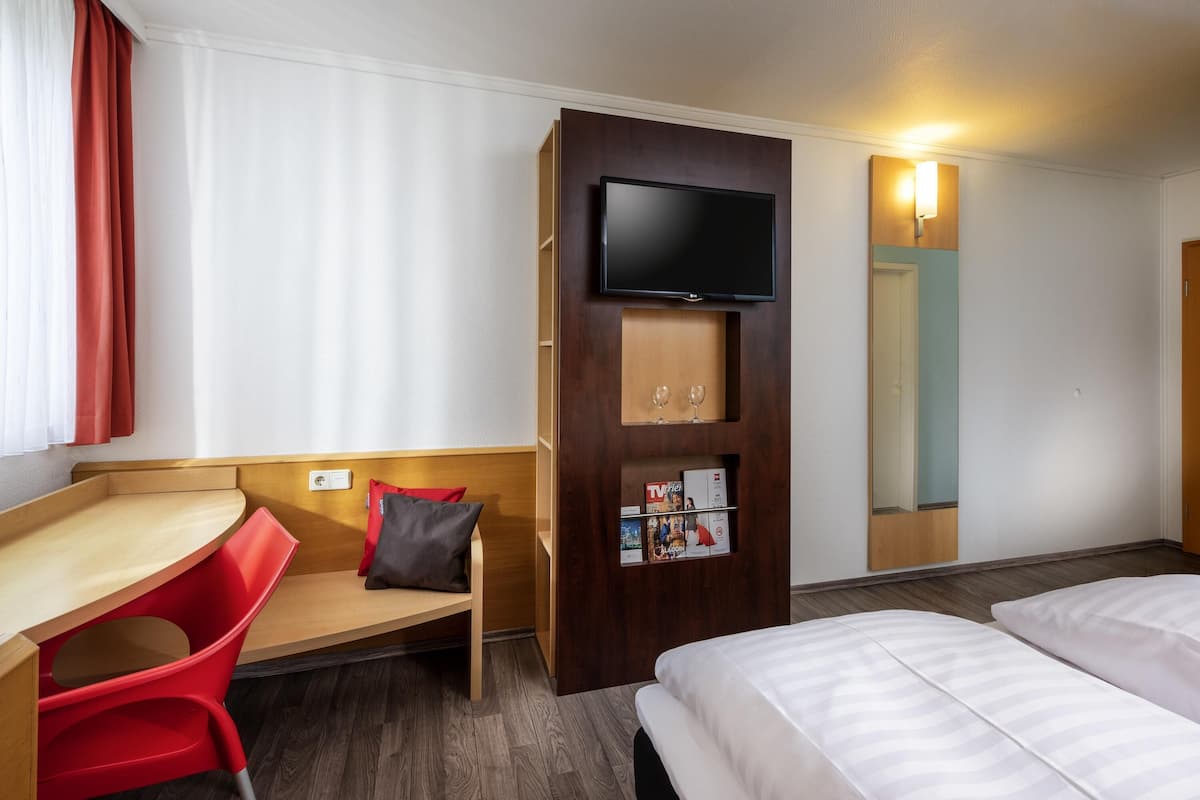 Ein Hotelzimmer mit Bett, Schreibtisch mit Stuhl, einem an einer Holzvertäfelung befestigten Fernseher und einem Spiegel an der Wand.