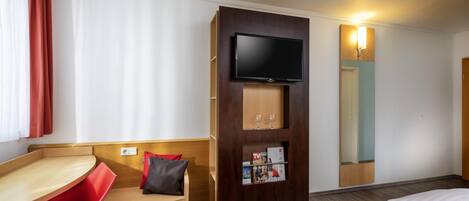 A hotel room with a bed, a desk with a chair, a TV mounted on a wooden panel, and a mirror on the wall.