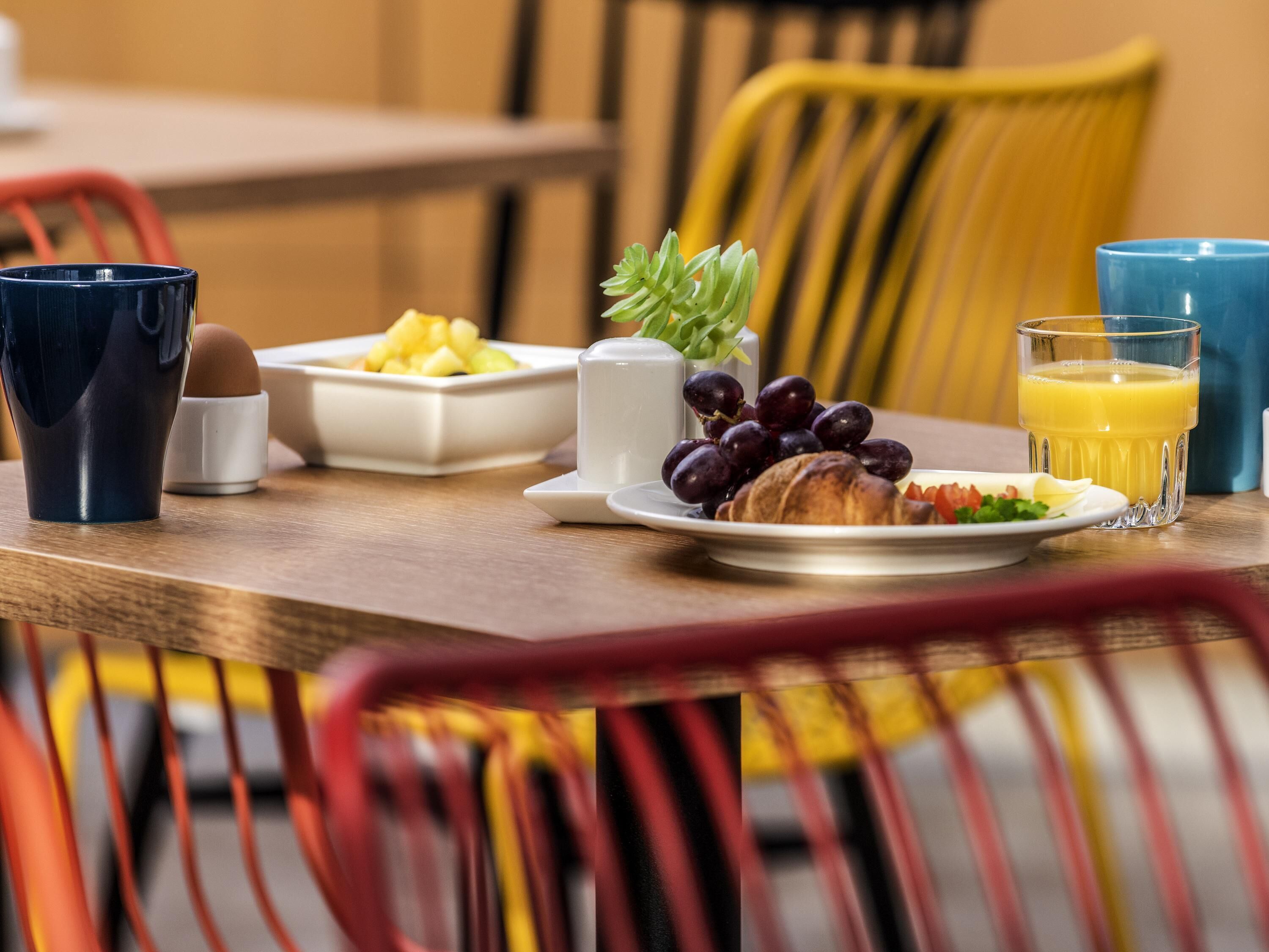A breakfast table with a croissant, grapes, a glass of orange juice, a blue mug, and a white cup.