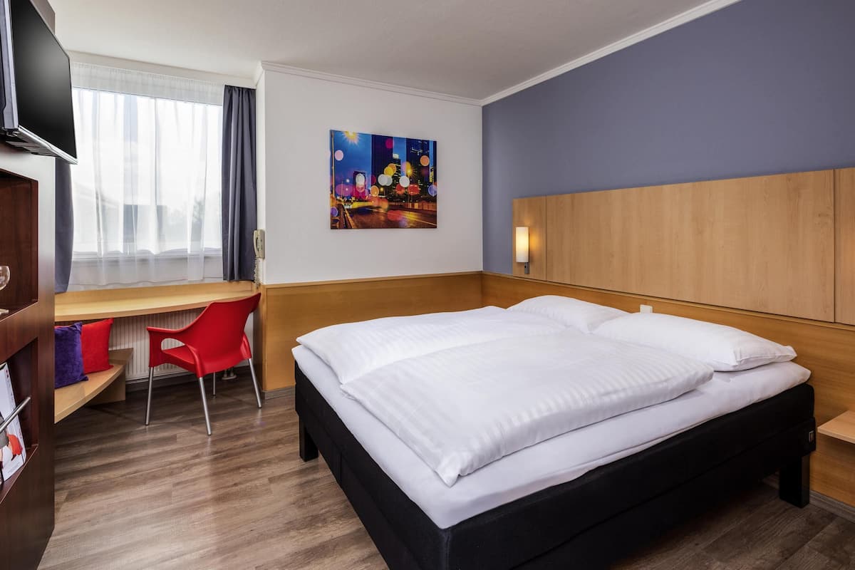A hotel room with a bed, a desk, a chair, a TV, and a painting on the wall.