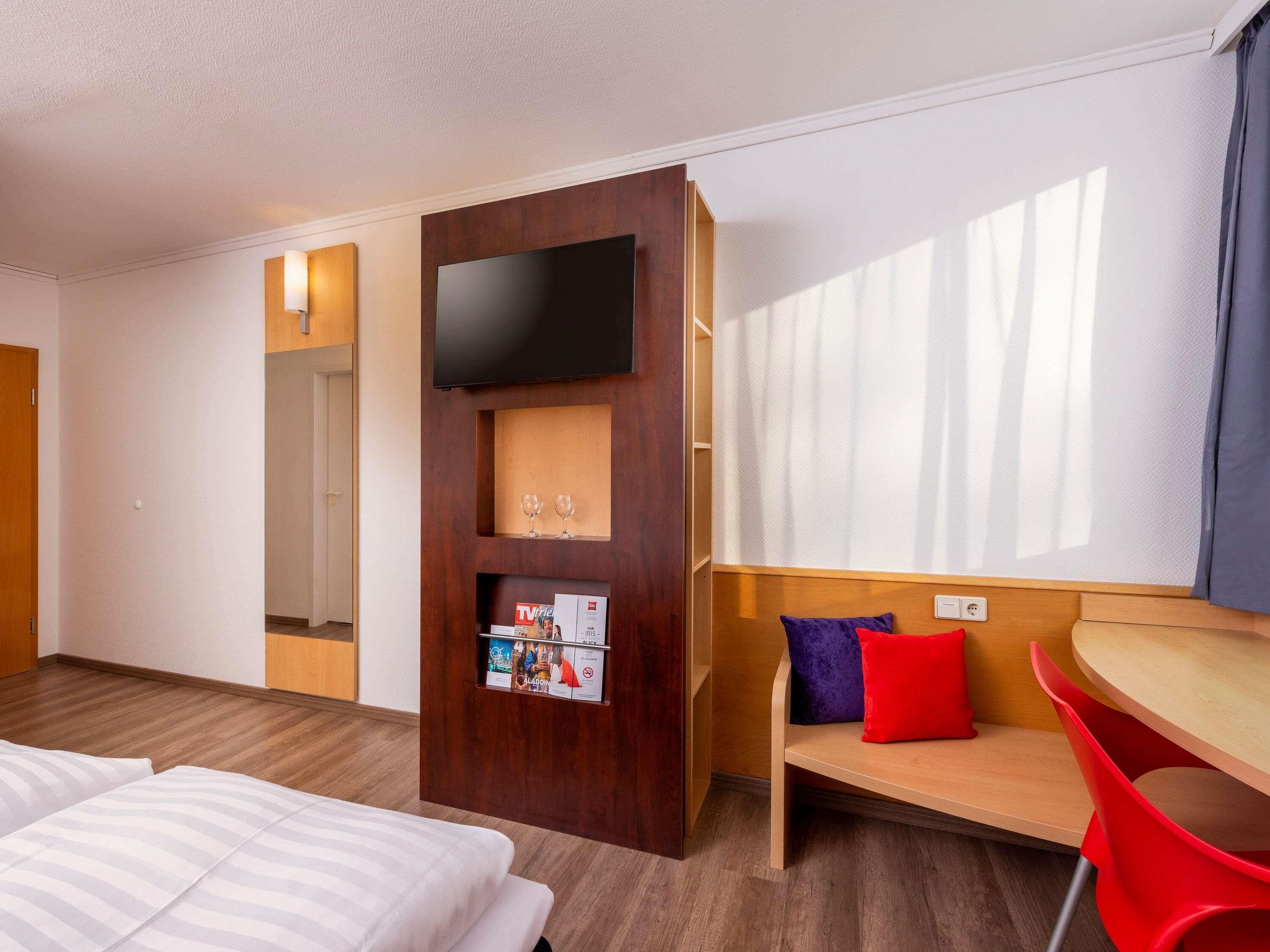A hotel room with a bed, a TV, a small table with a chair, and a window with curtains.