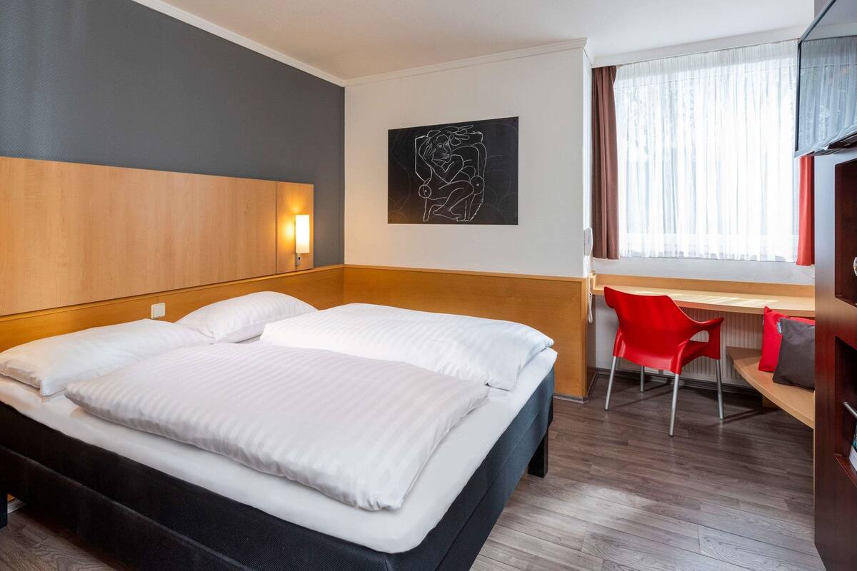 A hotel room with a large bed, a desk, a chair, and a window with curtains.