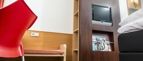 Habitación de hotel con un separador de madera, un televisor montado en él, una silla roja, un banco de madera y un sofá negro.