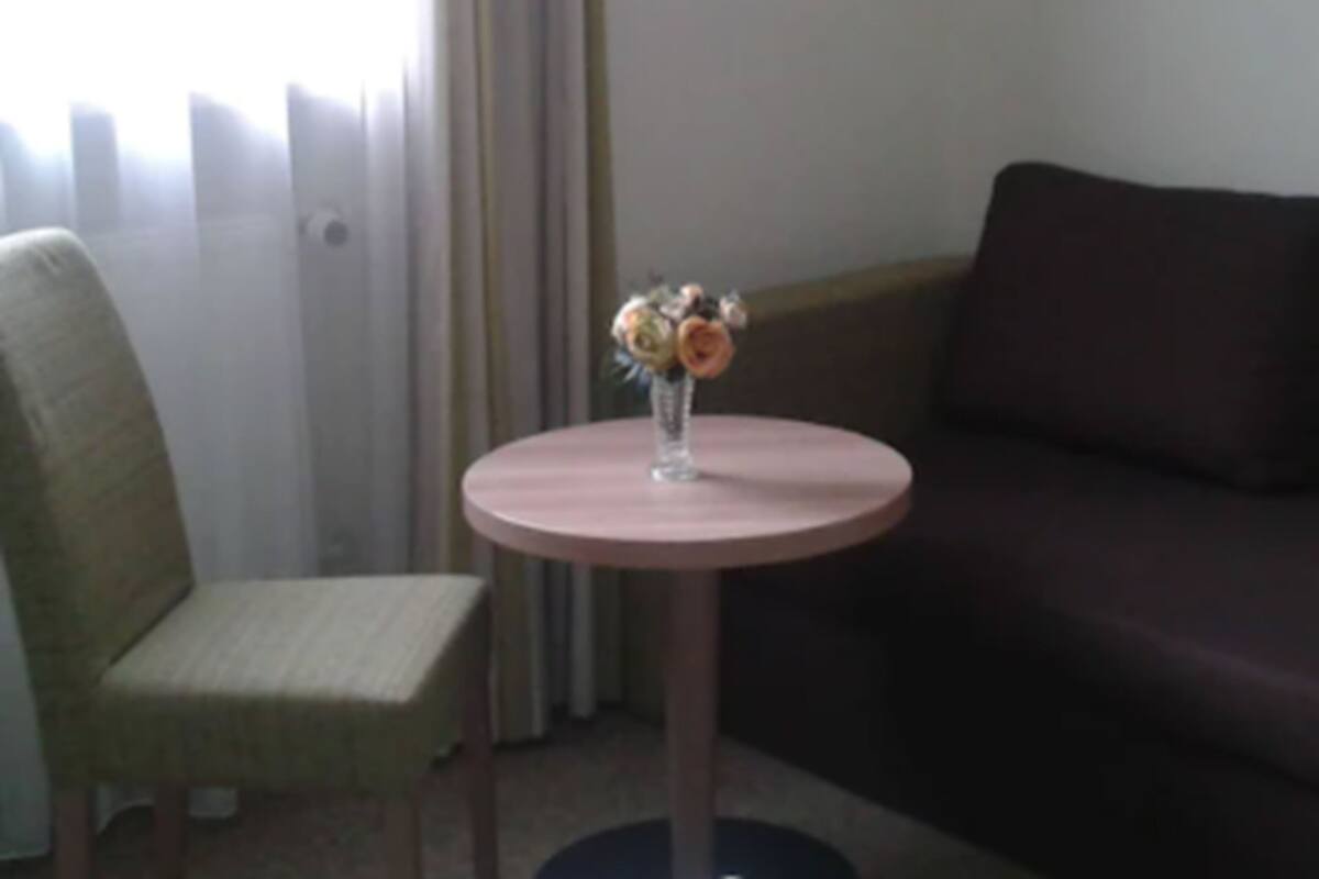 Una mesa pequeña y redonda con un florero, una silla verde y un sofá de color oscuro en una habitación con ventana y cortinas.
