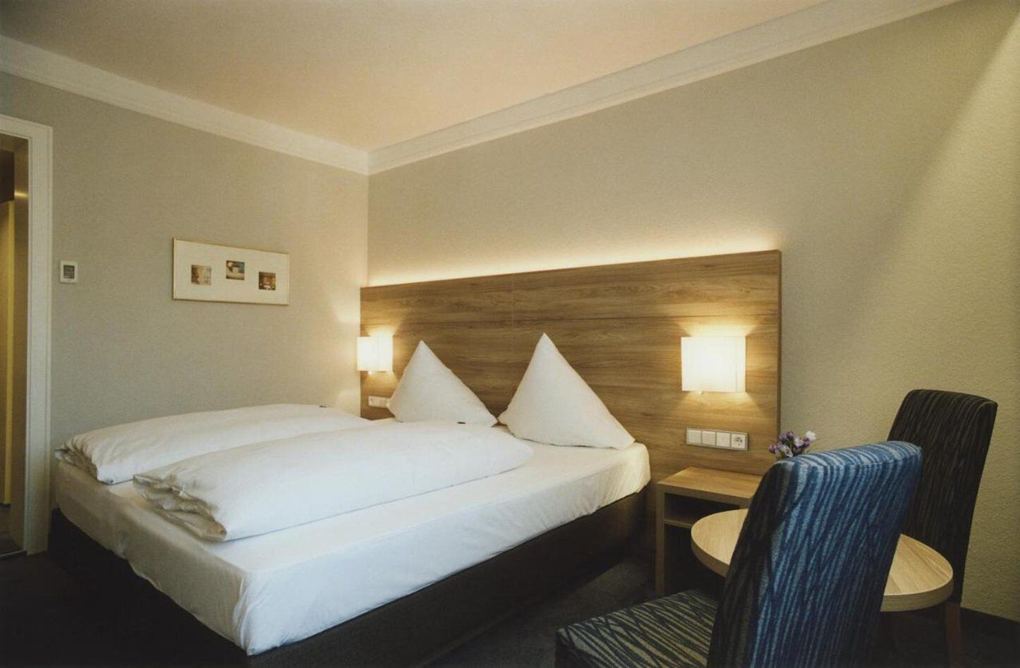 A hotel room with a double bed, two bedside tables with lamps, a small desk with a chair, and a framed picture on the wall.