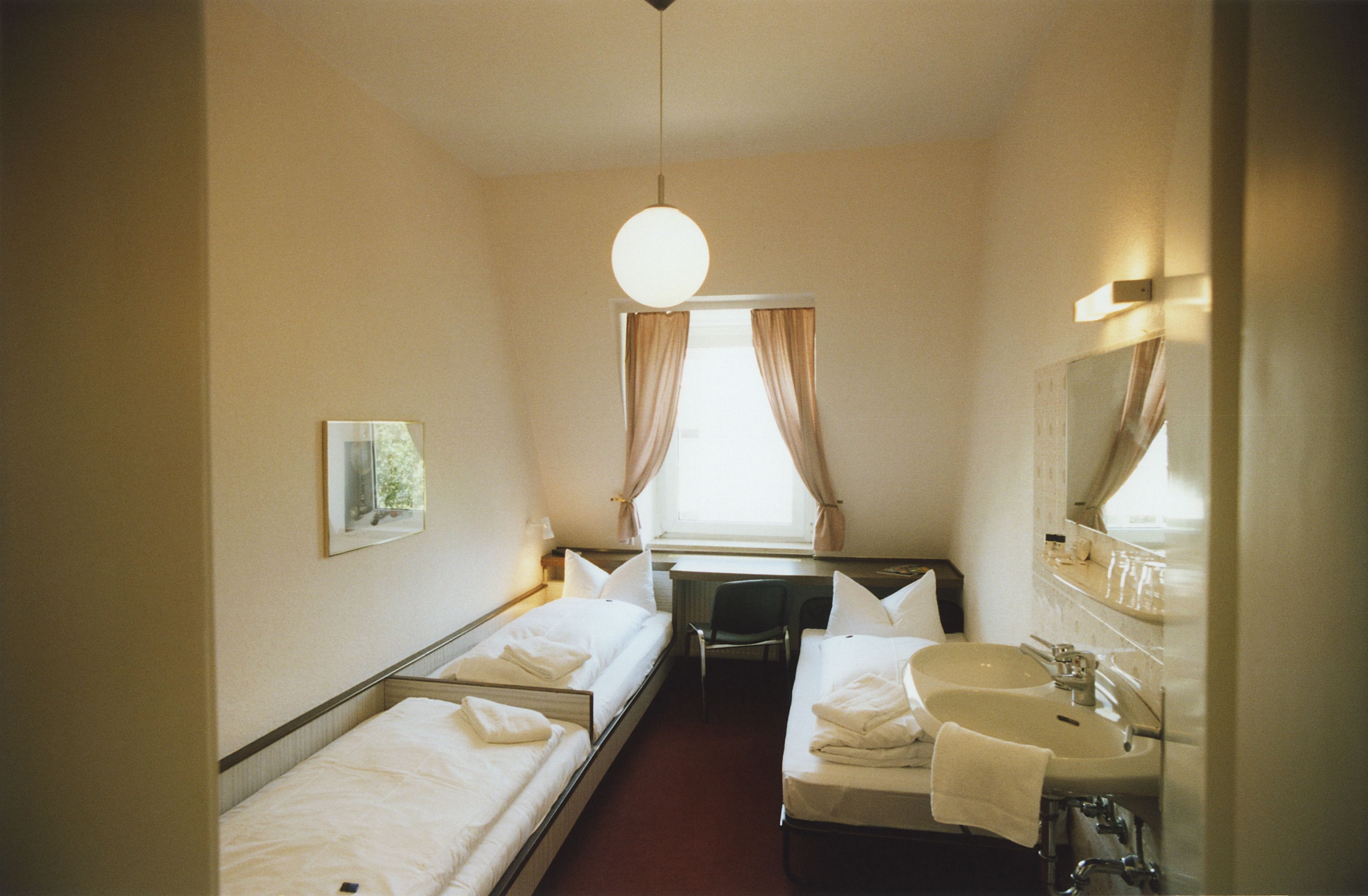 A hotel room with two single beds, a desk, a chair, a sink, and a mirror.