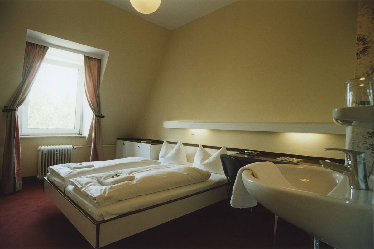 Habitación de hotel con cama, mesita de noche, lámpara, ventana con cortinas, radiador y baño con lavamanos y espejo.