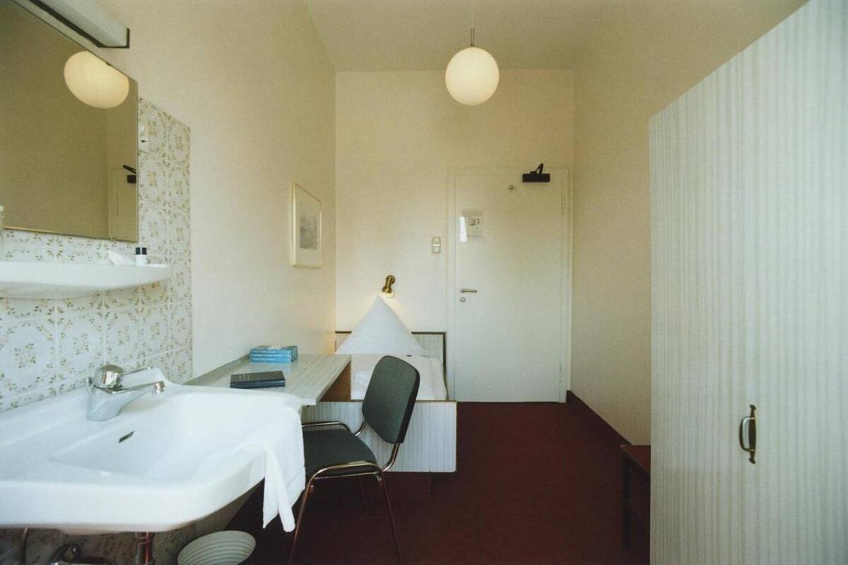Habitación de hotel pequeña y compacta con una cama individual, una mesita de noche, una silla, un escritorio y un espejo grande.