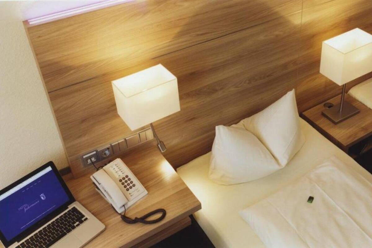 Habitación de hotel con cabecera de madera, dos mesitas de noche con lámparas, una computadora portátil, un teléfono y una cama bien hecha con almohadas.