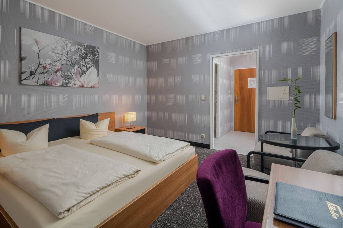 A hotel room with a large bed, a desk with a chair, a small table with a vase, and a door leading to another room.