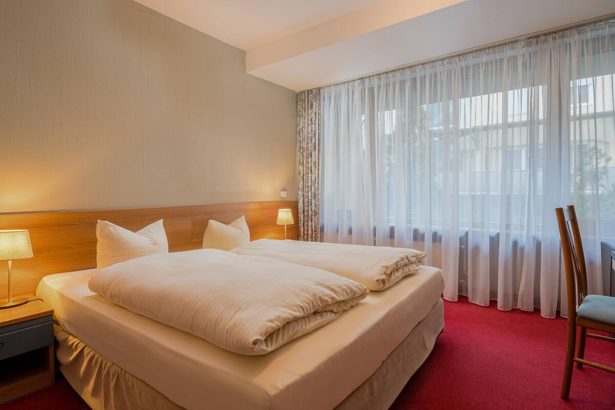 A hotel room with a large bed, two bedside tables with lamps, a chair, and a view through sheer curtains.