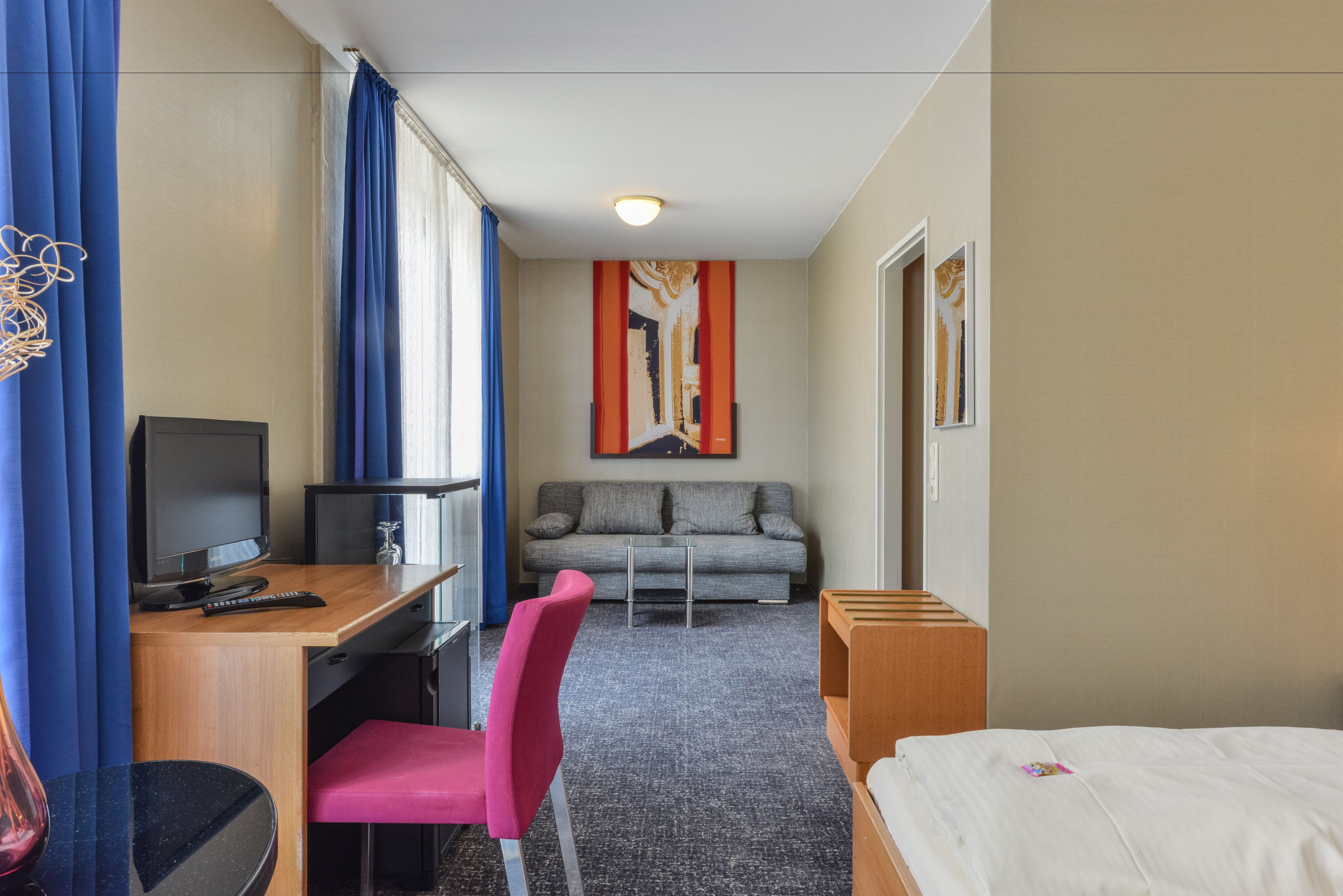 A hotel room with a bed, a desk with a computer, a chair, a sofa, and a TV.