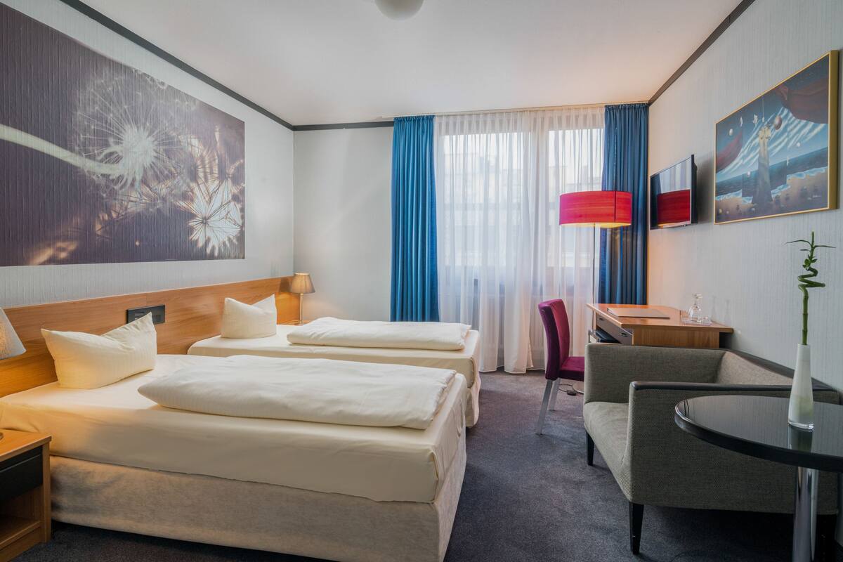 A hotel room with two beds, a desk, a chair, and a large window with curtains.