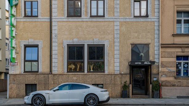 Ein weißes Auto vor einem Hotel mit einer steinernen Fassade und zahlreichen Fenstern.