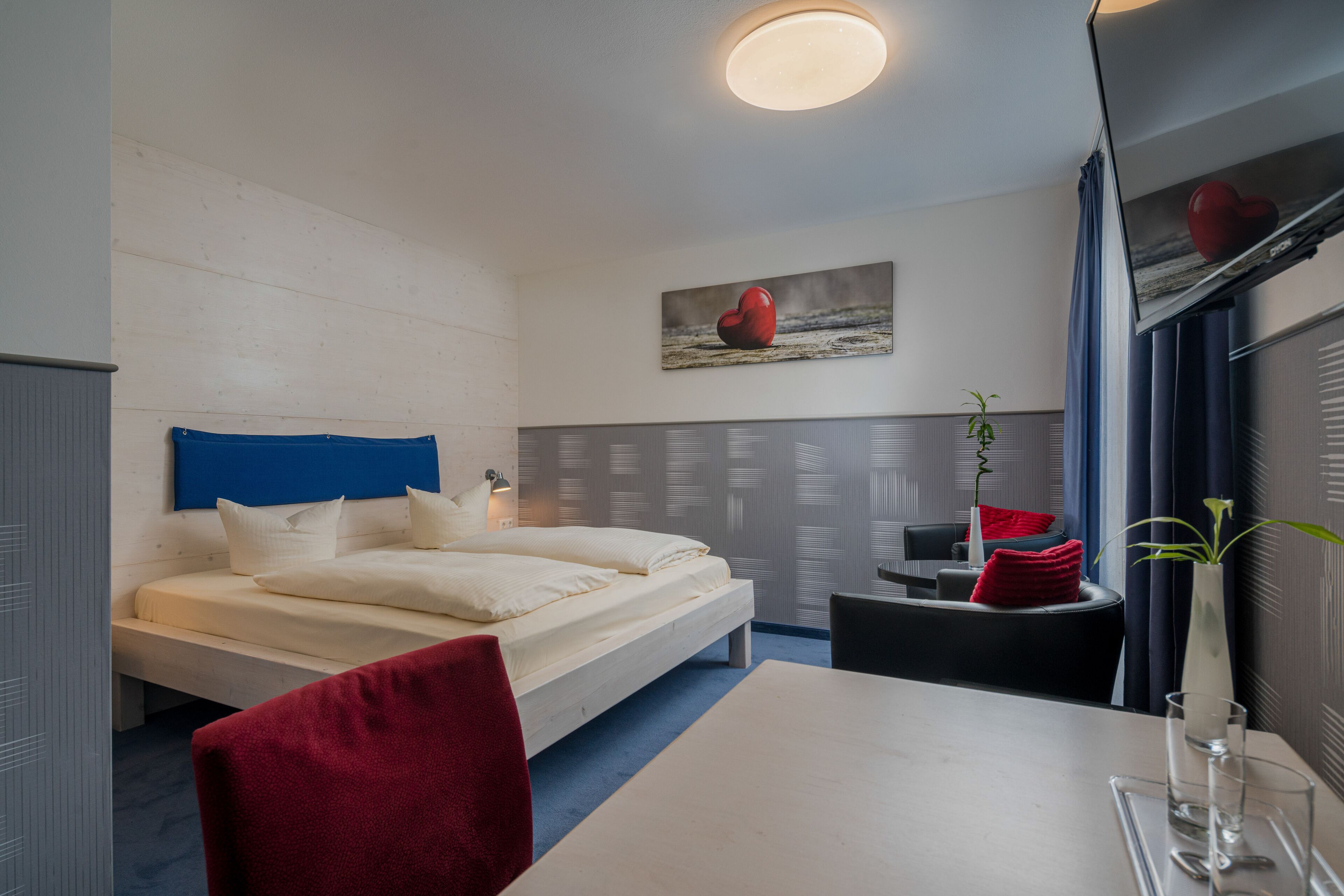 A modern hotel room with a bed, a dining table, a chair, a small table with a vase and glasses, and a painting of a heart on the wall.