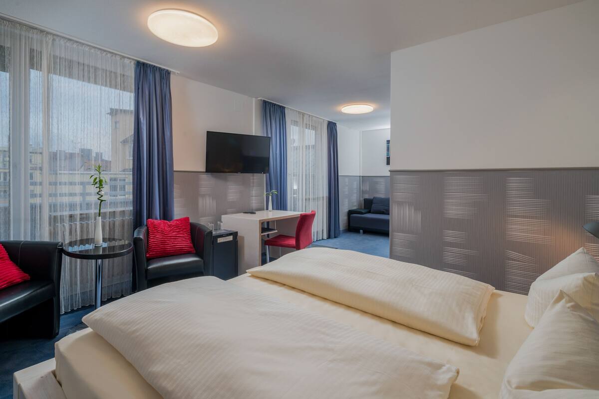 A hotel room with a large bed, a desk, a chair, a TV, and a view of the city.