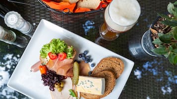 Ein weißes, rechteckiges Tablett mit verschiedenen Speisen, darunter Aufschnitt, Käse und Gemüse, dazu ein Glas Bier und ein Korb mit Brötchen.