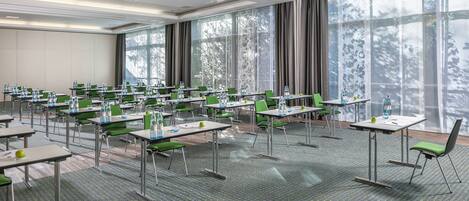 Uma sala de conferências com várias mesas e cadeiras, grandes janelas com cortinas e piso coberto de carpete.