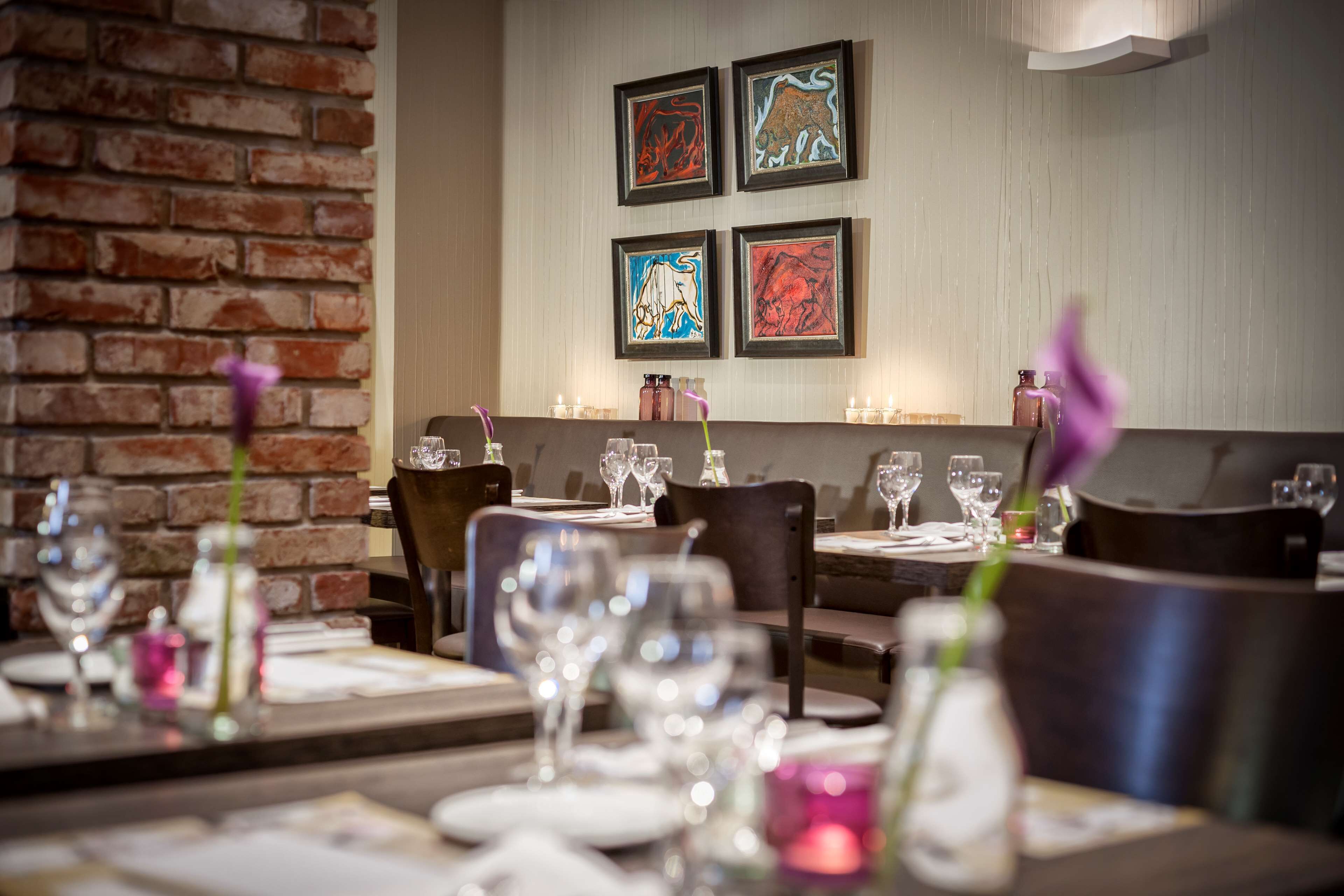 A restaurant interior with brick walls, framed artwork on the wall, and tables set with glasses and cutlery.