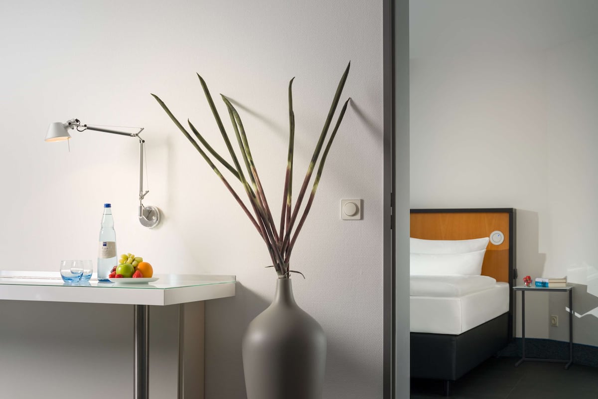 Una habitación de hotel con cama, mesita de noche, lámpara, jarra con plantas y frutero.
