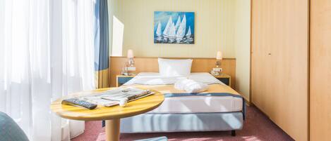 A hotel room with a bed, a small table with a remote control, a chair, and a painting on the wall.