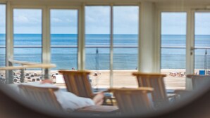 Habitación con grandes ventanales que dan a una playa. Se ven varias sillas de madera y una mesa en primer plano.