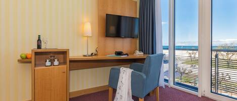 A hotel room with a wooden desk, a television mounted on the wall, a chair, a small cabinet with bottles, and a view of the sea through large windows.