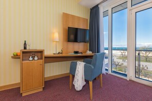 A hotel room with a wooden desk, a television mounted on the wall, a chair, a small cabinet with bottles, and a view of the sea through large windows.