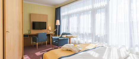 Habitación de hotel con cama, escritorio, silla, televisión y ventanas grandes con cortinas.
