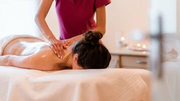 A massage therapy session in a spa setting with a client lying face down on a treatment table, a therapist attending to the client, and a small table with candles in the background.