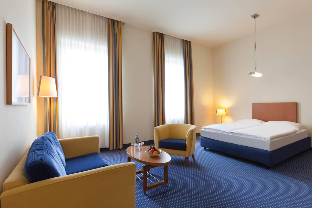 A hotel room with a bed, two chairs, a small table with a fruit bowl, a lamp, and a mirror.