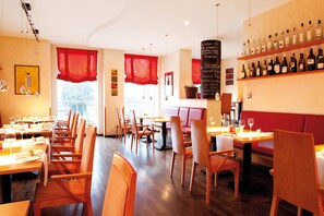 Interior de un restaurante con mesas y sillas de madera, una estantería para vinos en la pared y un banco con cojines rojos.