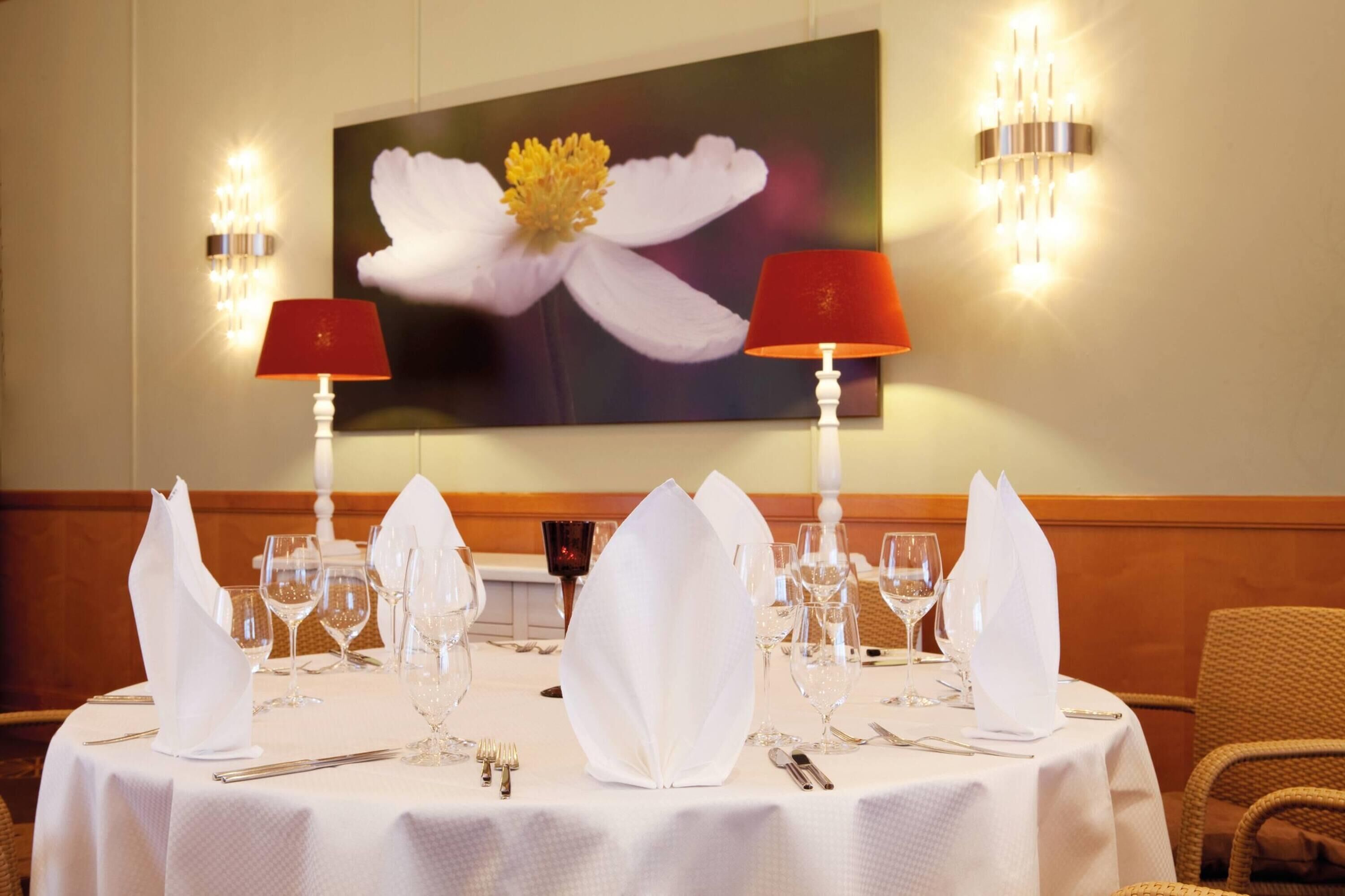 Una mesa de comedor dispuesta con servilletas blancas, copas de vino y cubiertos, con un cuadro grande de flores en la pared de fondo.