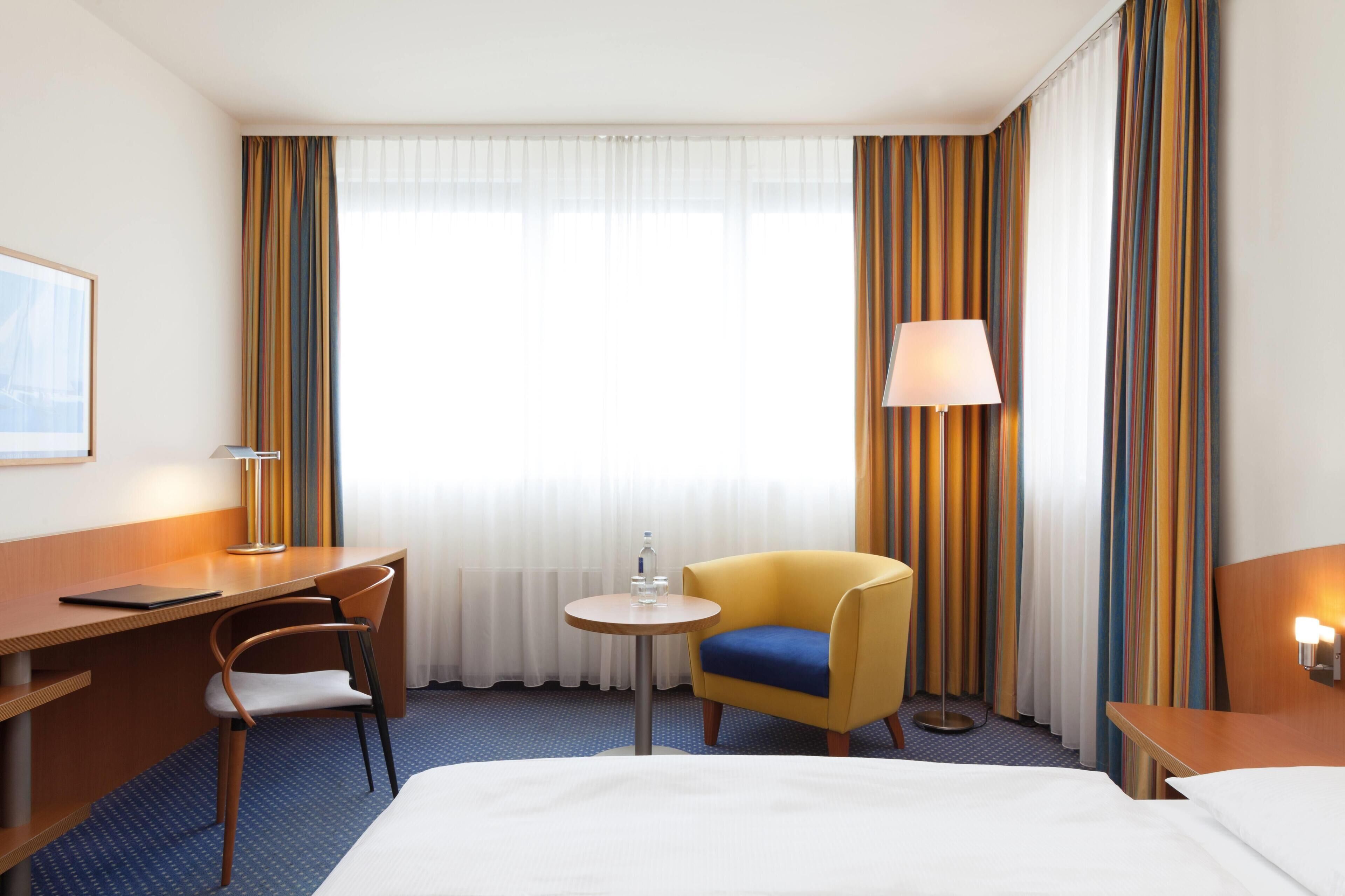 A hotel room with a bed, a desk, a chair, a small table, a lamp, and curtains.