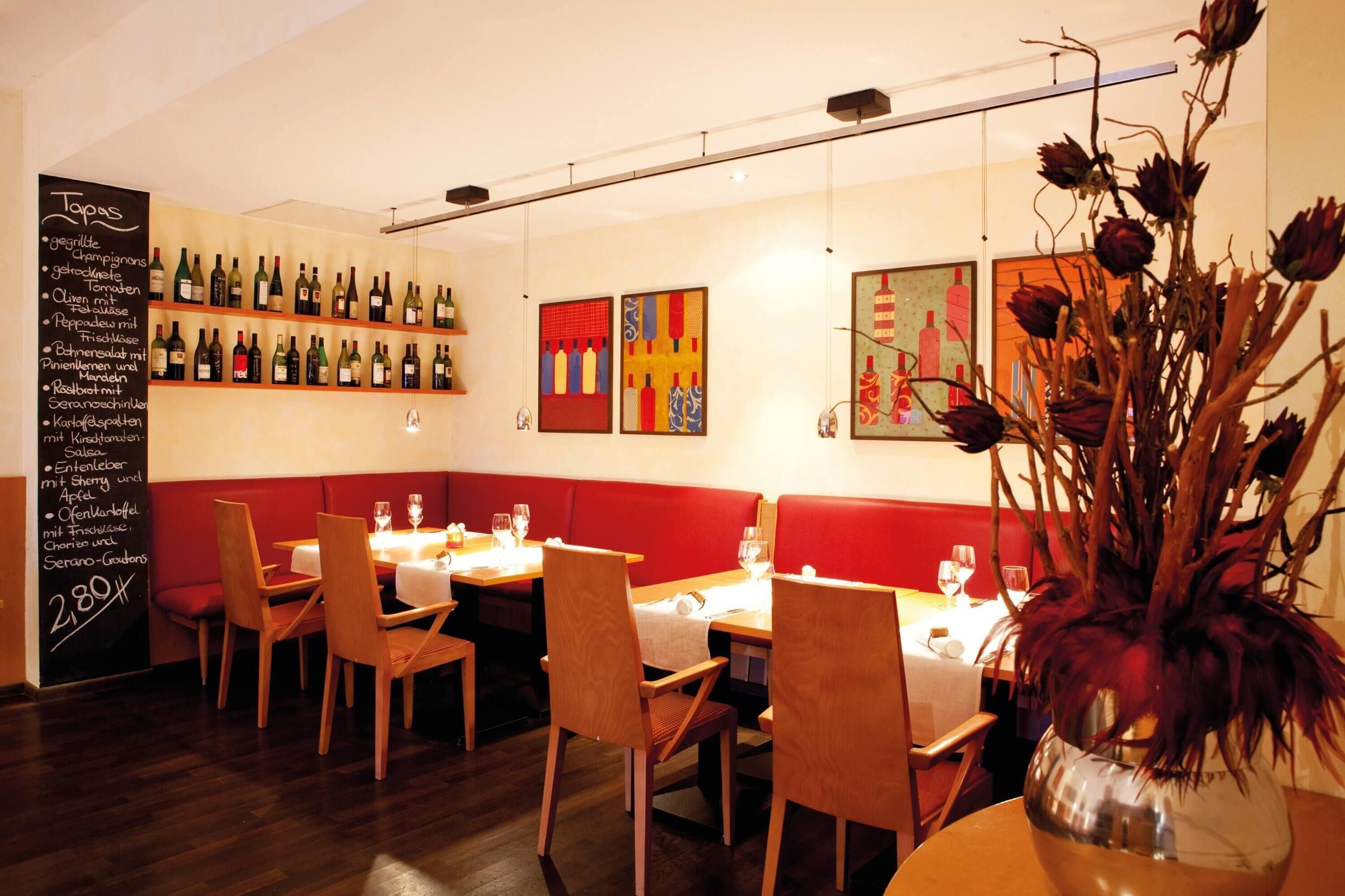 A restaurant interior with wooden chairs, tables, a chalkboard menu, and wine bottles on shelves.