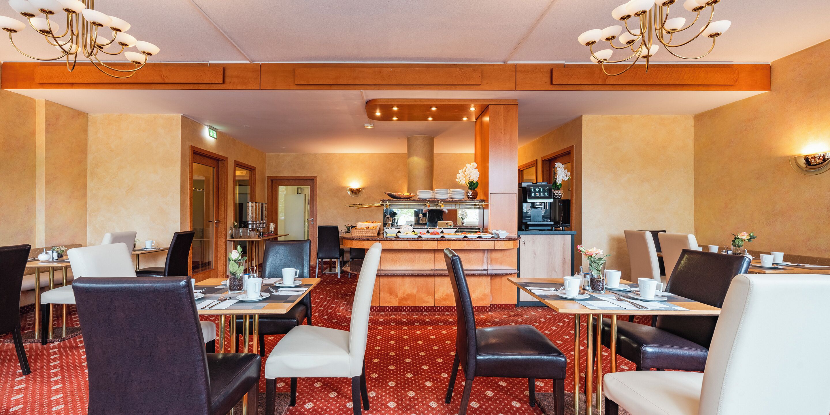 Comedor de hotel con mesas y sillas, una cocina al fondo y un candelabro.