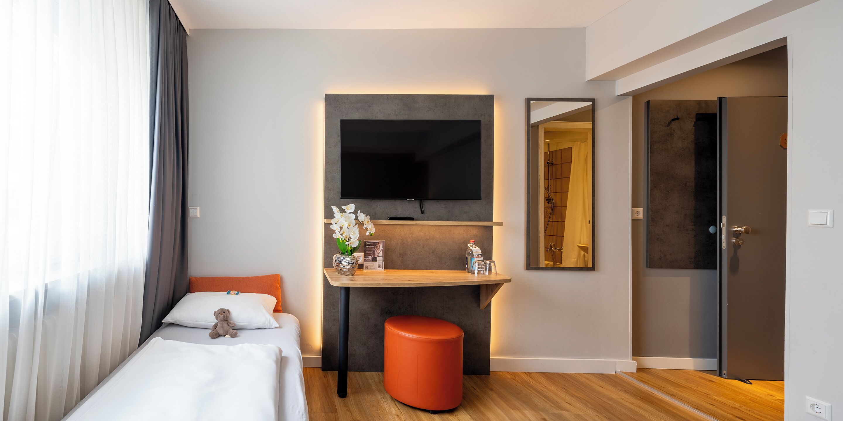 A modern hotel room with a bed, a small table with a vase of flowers, a mirror, and a television mounted on the wall.