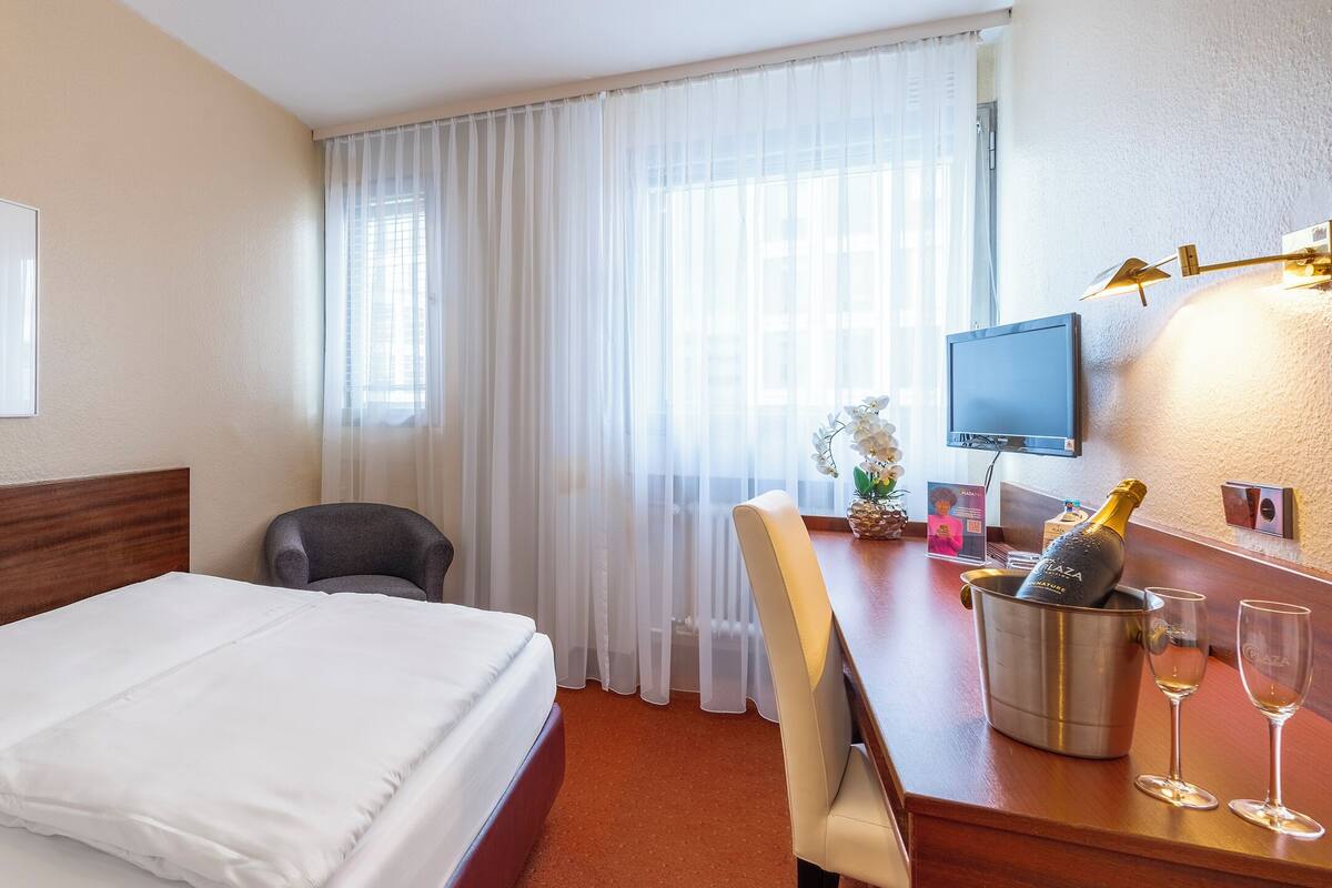 A hotel room with a bed, a desk with a computer, a chair, a window with curtains, and a framed picture on the wall.